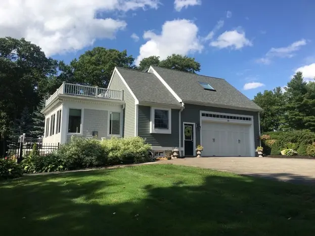 A modern gray and white house with Vinyl Siding Installation in Newton, MA, featuring a two-car garage, small upper balcony, and large windows. The landscaped front yard has green grass and shrubs, framed by trees and a blue sky with scattered clouds.