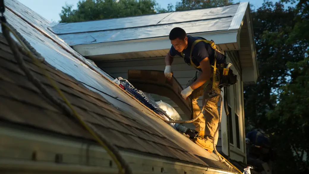 A construction worker wearing safety gear installs shingles on a sloped house roof during daylight, surrounded by trees, while exterior remodeling continues with another worker partially visible in the background.