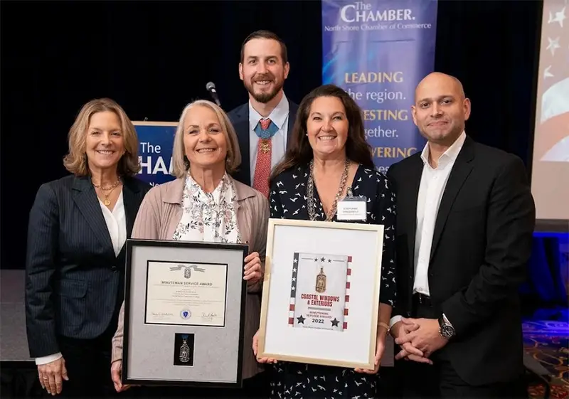 Five people stand together at the North Shore Chamber Salute to Veterans Breakfast, smiling. Two women in the center hold framed awards. A sign behind them reads “The Chamber, North Shore Chamber of Commerce.” A podium is on the left.