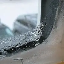 Close-up of an insulated window corner with visible condensation and water droplets on the glass. The outer scene is blurred, suggesting the window is part of a building facing a street or parking lot. Curtains add to the overcast mood, enhancing the damp atmosphere.
