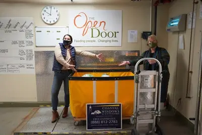 Two people wearing masks stand beside a large yellow food bin at The Open Door food pantry. One gestures toward the bin; the other holds a cart. A Coastal Windows & Exteriors In The News Submission #5 sign and clock are visible on the wall behind them.