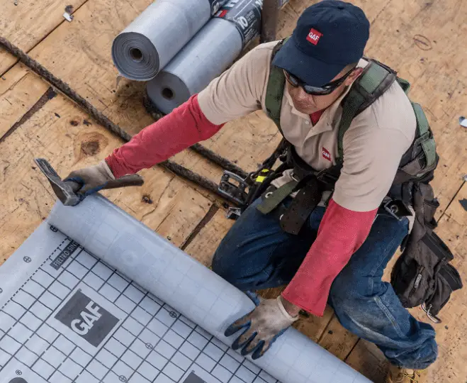 A construction worker in safety gear installs roofing underlayment for roof deck protection on a wooden roof, using a hammer. Rolls of underlayment and tools are visible nearby. The worker wears a GAF-branded cap and shirt.