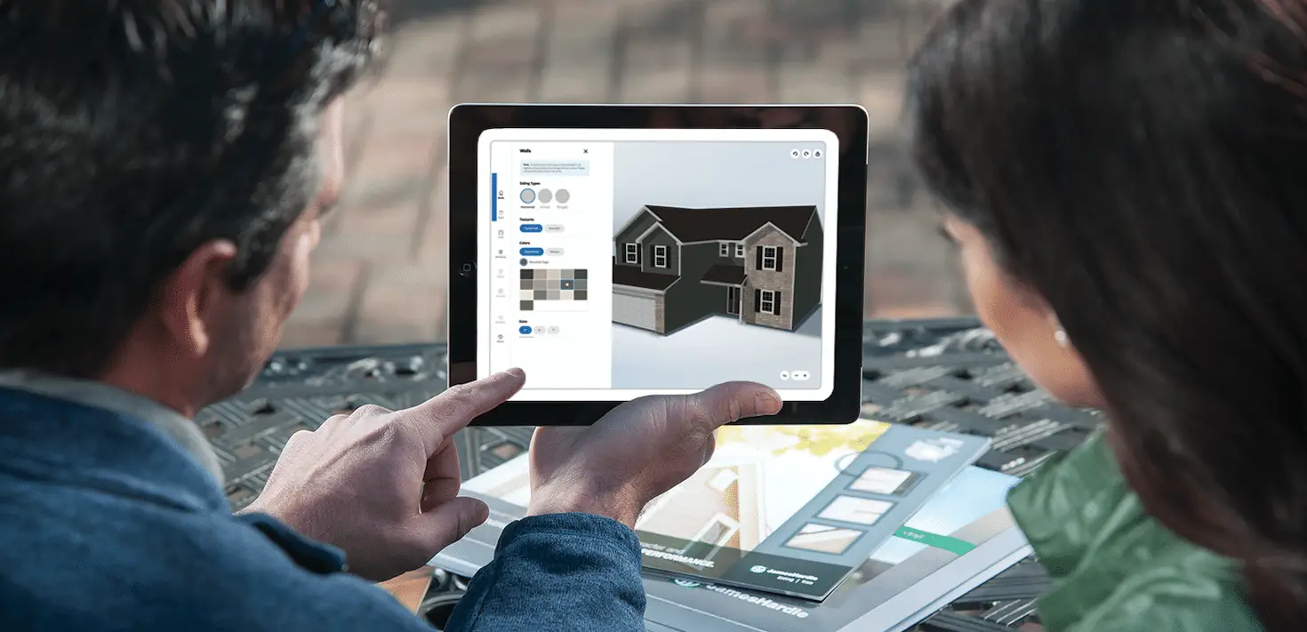 Two people look at a tablet displaying a 3D model of a house, exploring options for windows and siding. Sitting outside with brochures in front of them, they discuss home design possibilities, perhaps considering choices linked to a referral program focused on renovations.