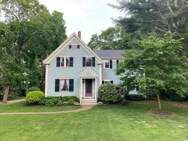 A two-story light blue house with dark shutters and a central front door, surrounded by green trees, shrubs, and a well-kept lawn—showcasing quality roofing in Waltham MA on a bright, sunny day.