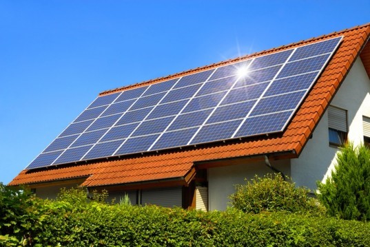 A house with a red-tiled roof meets all roof requirements for solar panels, which are installed on top and reflect sunlight under a clear blue sky. Green shrubs border the house in the foreground.