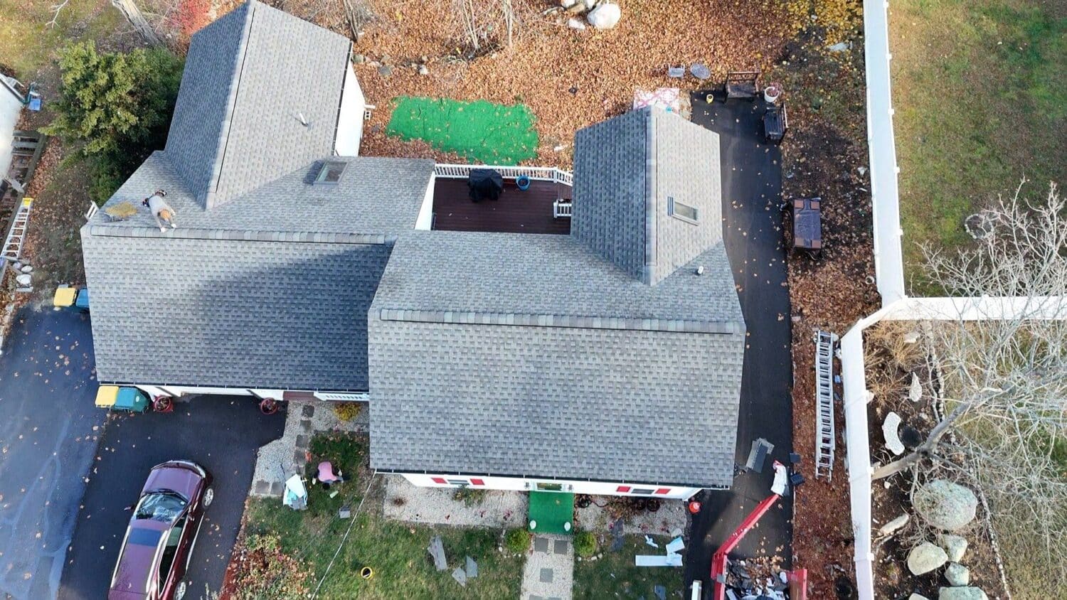 Aerial view of a house with a gray shingle roof surrounded by autumn leaves, people working on the roof and yard, a red vehicle in the driveway, and equipment scattered around the property.