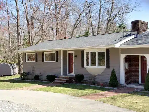Single-story house with beige siding, white trim, dark shutters, and a red front door; a small porch with steps leads to the entrance. There is a carport on the right and a shed in the background among leafless trees—ideal for spring roofing updates.