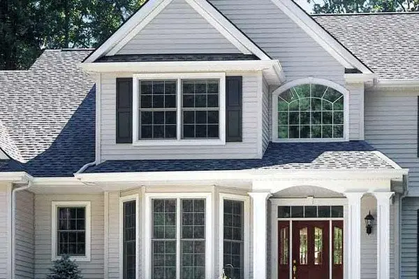 Two-story house with light gray siding, dark gray roof, and white trim. Perfect for summer home renovations, it features large front windows, including a bay window and arched window, plus a covered entryway and double red front doors.