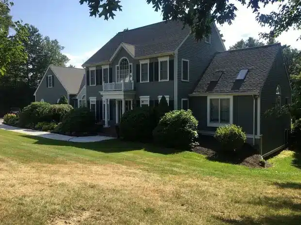 A two-story dark gray house with white trim features large windows and a balcony above the front entrance, showcasing expert Vinyl Siding Replacement in Brookline MA, all surrounded by shrubs and a well-maintained lawn on a sunny day.