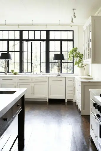 Bright, modern kitchen with white cabinets, dark wood floors, black frame replacement windows, two black lamps by the sink, and green plants on the counter.