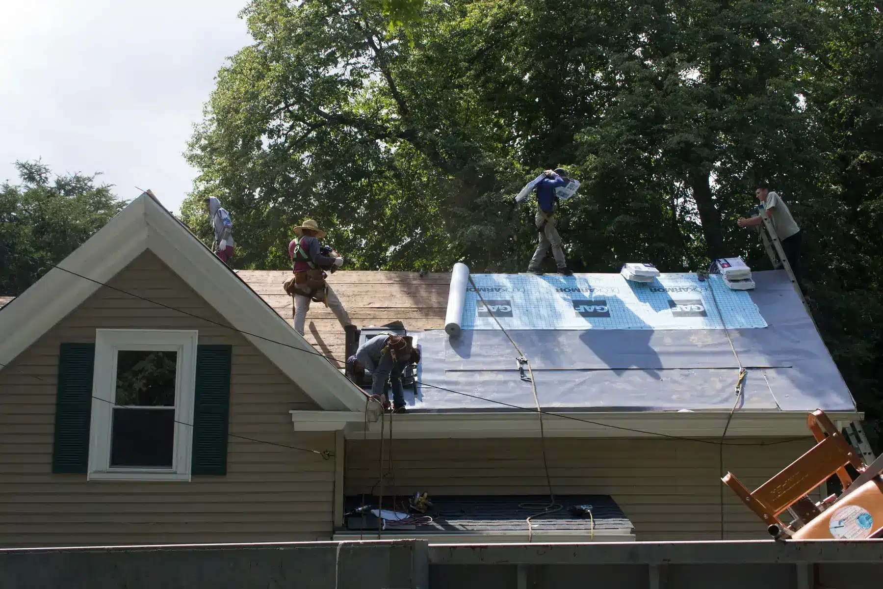Four workers are installing new roofing material with leak barrier protection on a house roof, using tools and supplies scattered around them. The house is surrounded by trees, and the workers are wearing safety harnesses.