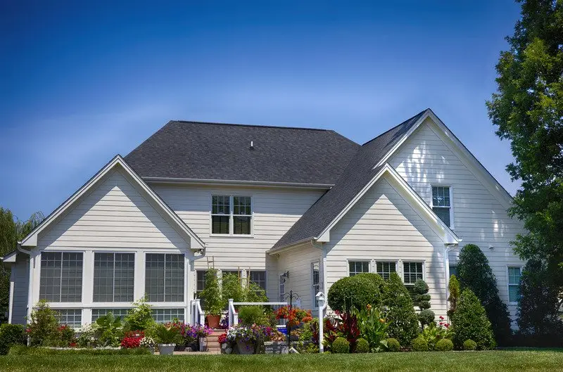 A modern white house with a dark roof, large windows—ideal if you’re wondering what is the best time of year to replace windows—and a lush garden filled with colorful flowers and neatly trimmed bushes, set against a clear blue sky.