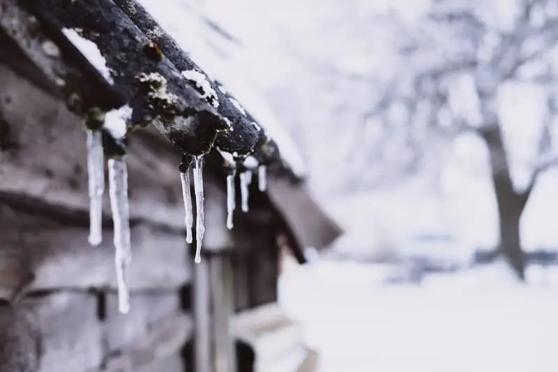 Icicles hang from the edge of a snow-dusted wooden roof—a familiar sight during winter roofing in New England—with a blurred, snowy landscape and bare tree in the background.