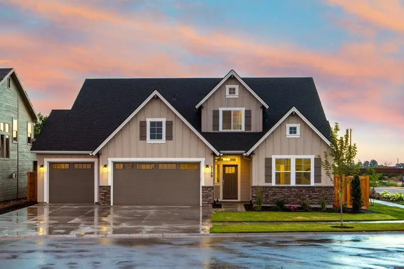A modern suburban house with beige siding, stone accents, and two garage doors glows warmly at sunset. Vinyl siding replacement can save you money while enhancing curb appeal, as seen in the wet driveway reflecting the colorful sky.