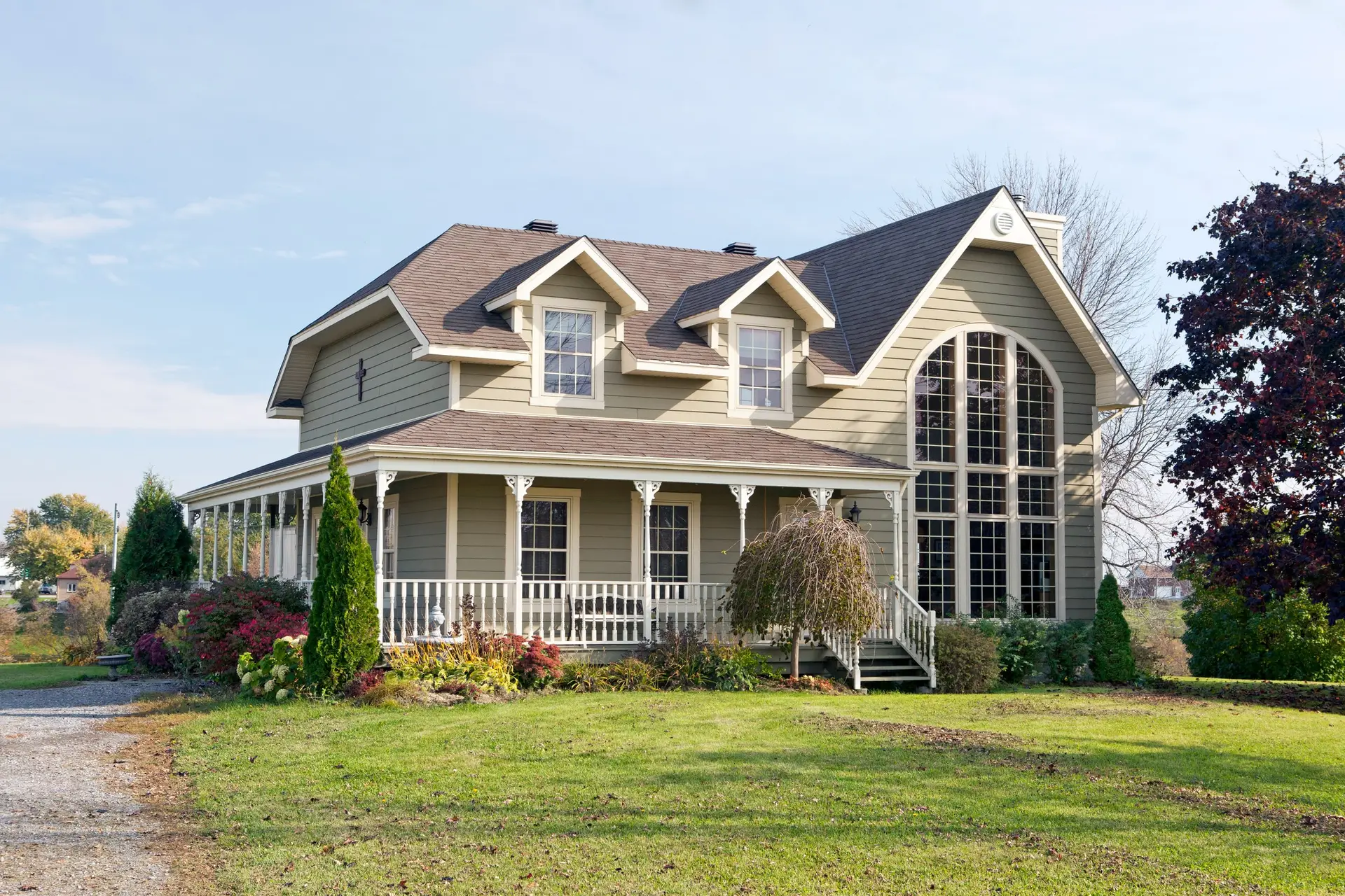 A two-story beige house with a large arched window, wraparound porch, and gabled roof sparkles under the blue sky, its decorative trim and vinyl siding gleaming—thanks to regular cleaning—amid green lawns, shrubs, and towering trees.