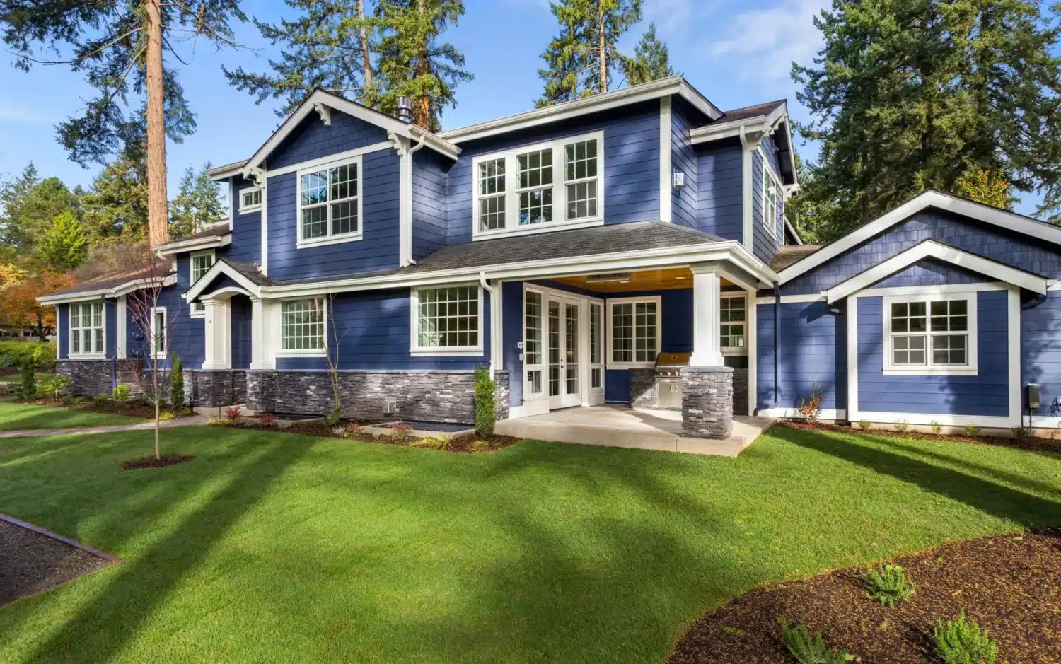 A large two-story blue house with white trim and stone accents, featuring many windows—perfect for using a Window Replacement Checklist—and a well-manicured lawn surrounded by tall trees under a clear blue sky.