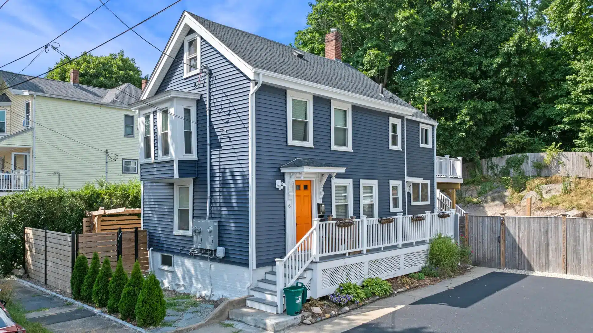 A two-story house with blue siding inspired by The Guide to Energy Efficient Siding, white trim, and an orange front door. It features a small front porch, potted plants, a paved driveway, and trees with neighboring homes in the background.