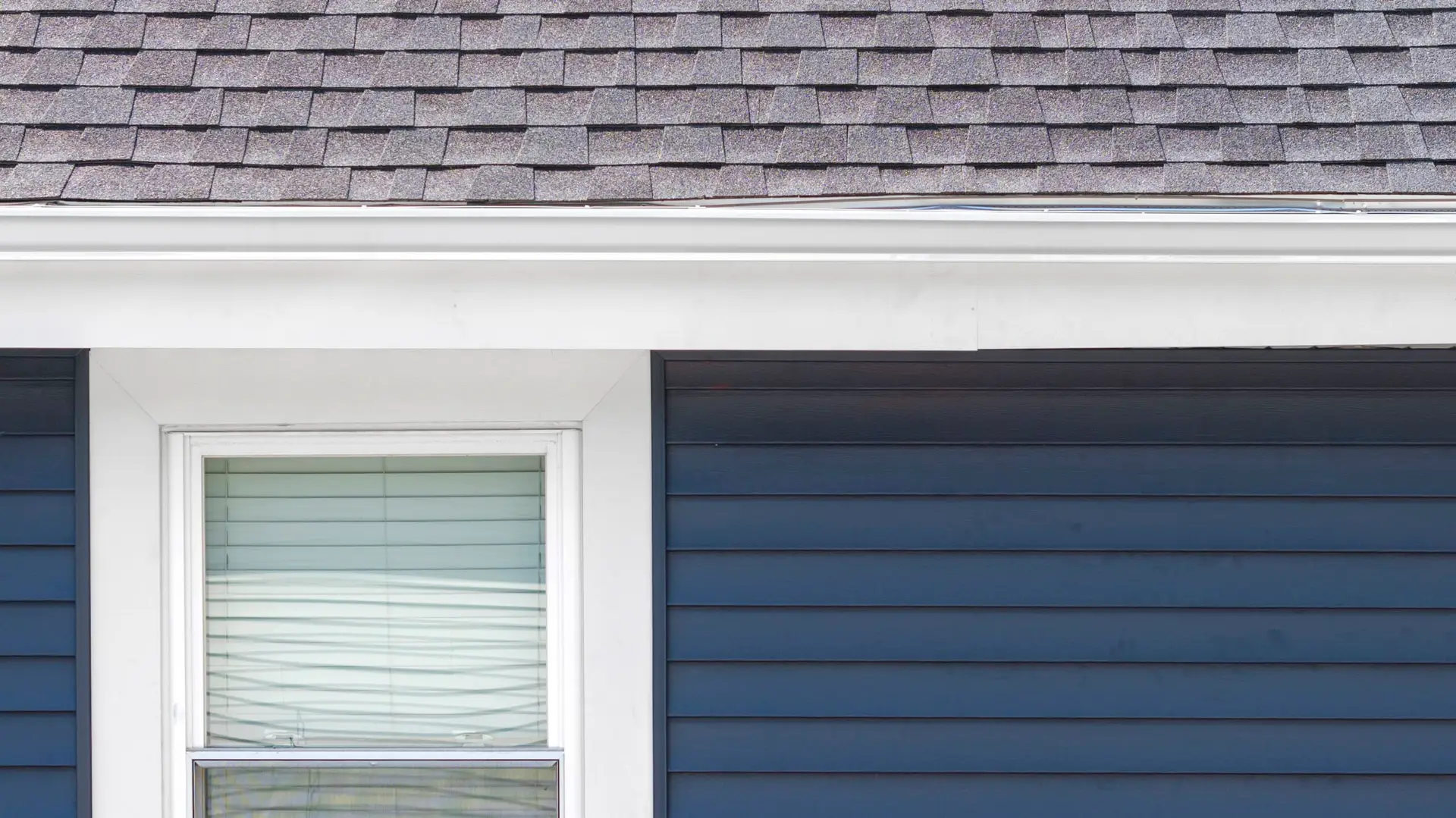 Close-up of a house exterior in Essex County featuring dark blue vinyl siding, a white-framed window with closed blinds, a white gutter, and a shingled roof.