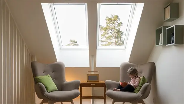 A young child sits on a gray armchair, reading a book in a cozy attic room with two large skylights, another empty armchair, a small table, and trees visible outside—a perfect setting to enjoy after a recent roof replacement.