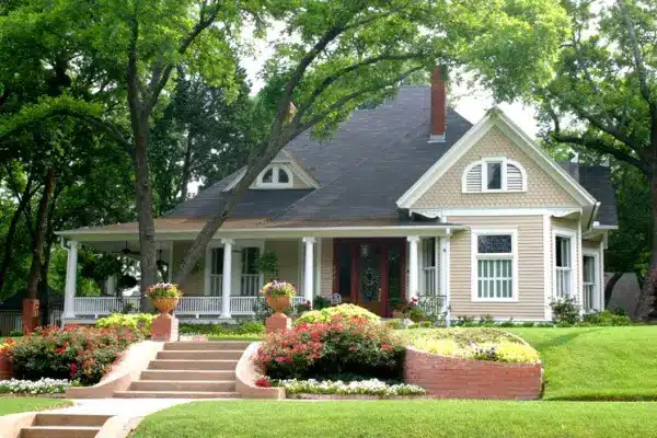 A charming two-story house with beige siding showcases recent exterior home improvement, featuring a large porch and steep roof among tall trees, surrounded by manicured lawns, colorful flower beds, and brick planters on a sunny day.