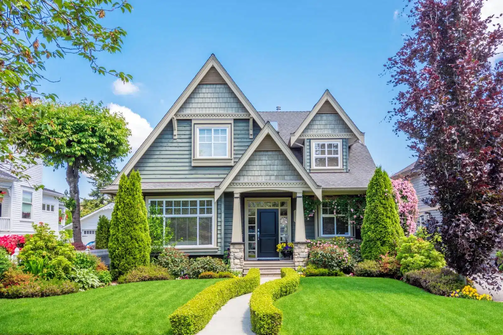 A charming two-story suburban house with gray siding, a gabled roof, and white trim is surrounded by lush greenery and colorful flower beds—perfect for tackling spring home improvement tasks on a sunny day.