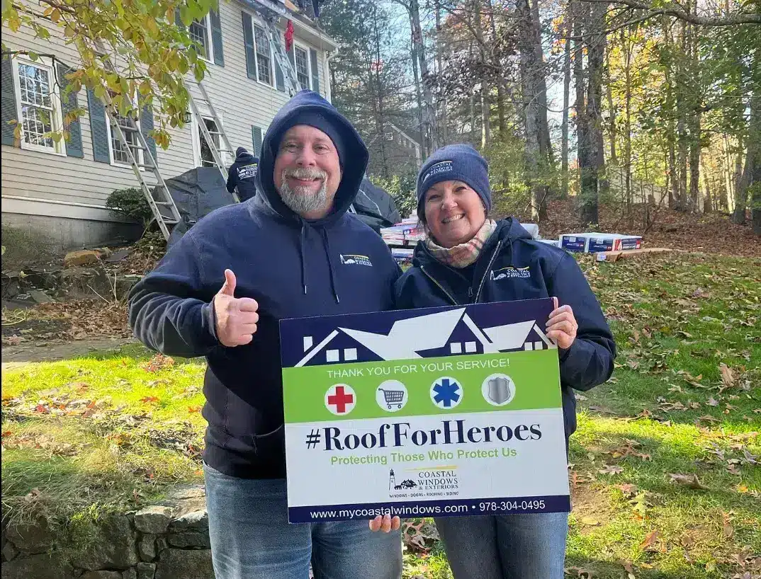 Two people in jackets and beanies smile outdoors, giving thumbs up and holding a #RoofForHeroes sign by Roofs For Heroes - My Coastal Windows, standing in front of a house with workers and roofing materials.