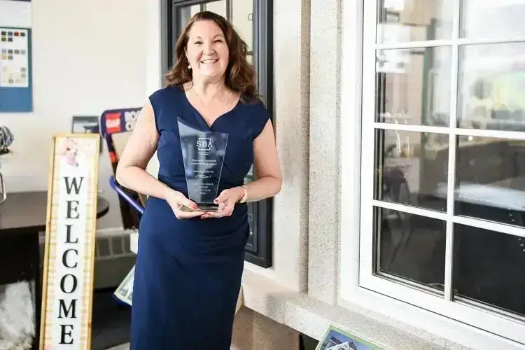 A smiling Beverly Woman in a blue dress holds a Business Person of the Year award while standing indoors near a window. A Welcome sign is visible in the background, as captured by Salem News.