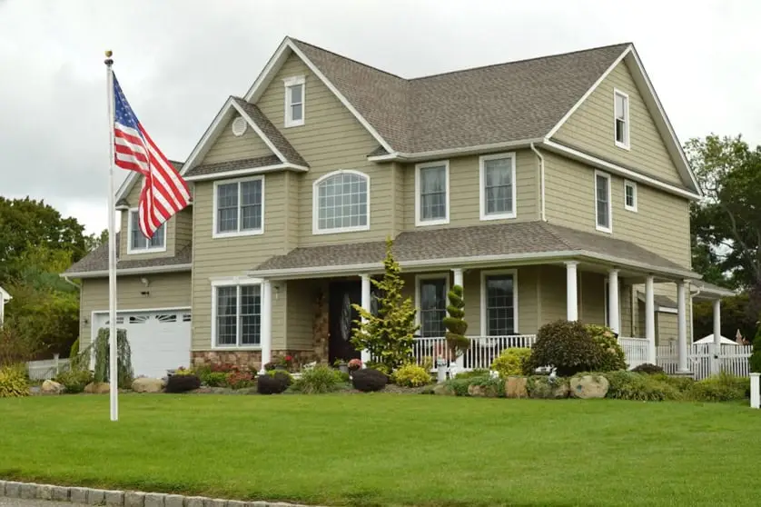 A two-story beige house with white trim features a large front porch and a well-manicured lawn with shrubs. An American flag stands in the yard under an overcast sky—perfect inspiration for your summer home improvement ideas.