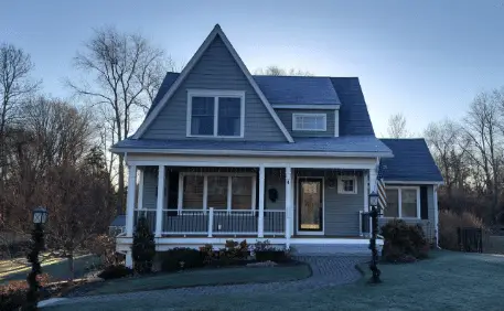A gray two-story house with white trim and a covered front porch sits on a grassy lawn. Bare trees and soft sunlight suggest a cool, early morning—highlighting the benefits of the engineering of energy-efficient windows.
