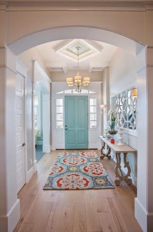 A bright entryway features a vintage door painted light blue, a colorful patterned rug, a wooden console table with decor, a mirror on the wall, and a chandelier on the tray ceiling. Light wood floors and white walls add to the dreamy, airy feel.