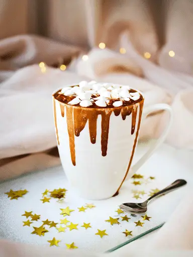 A white mug of hot chocolate topped with marshmallows and chocolate drips, set on a tray with gold star confetti and a spoon—perfect for cozy moments during our special Cyber Week Sale. Soft beige fabric and fairy lights glow in the background.
