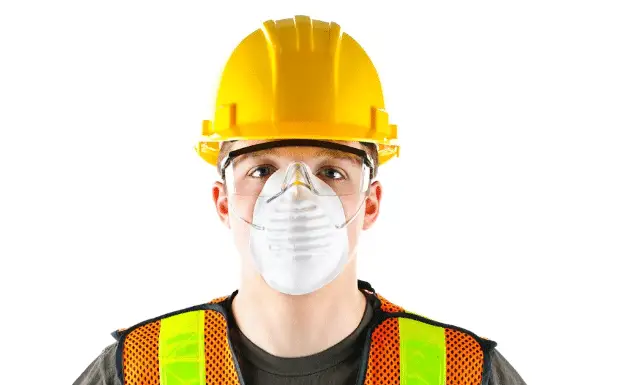 A construction worker wearing a yellow hard hat, safety glasses, a high-visibility vest, and a protective face mask stands against a white background, ready to work on exteriors for Coastal Windows projects.