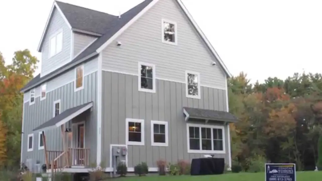 A modern, three-story gray Net-Zero Energy Home with white trim sits on a grassy lawn dotted with trees. Multiple windows brighten the space, and a small porch welcomes you up the steps. A sign is visible on the lawn for Wednesday Weekly Wow.