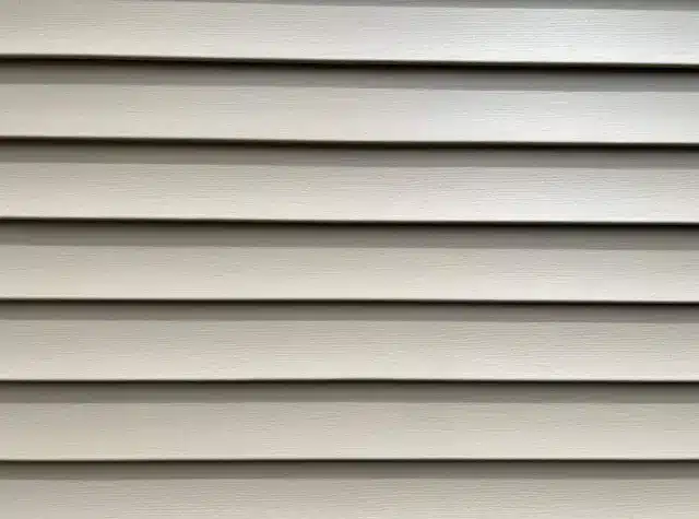 Close-up of beige horizontal vinyl siding panels, evenly spaced and parallel, covering an exterior wall. The smooth surface with subtle texture and shadows suggests a clean installation. For personalized style, one might wonder: can I paint vinyl siding to refresh its appearance?.