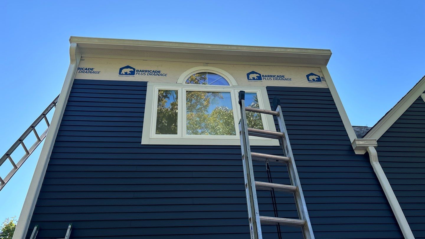 A house under construction with dark blue siding, white trim, and an arched window. Two ladders are leaned against the house, reaching towards the roof. Sections of the wall are covered with house wrap material. The sky is clear and blue.