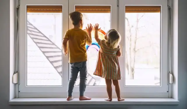 Two young children stand on a windowsill, looking outside. One wears a yellow shirt and pants, the other a striped dress. Both press their hands against the glass, daylight shining in—a gentle reminder during National Window Safety Week.