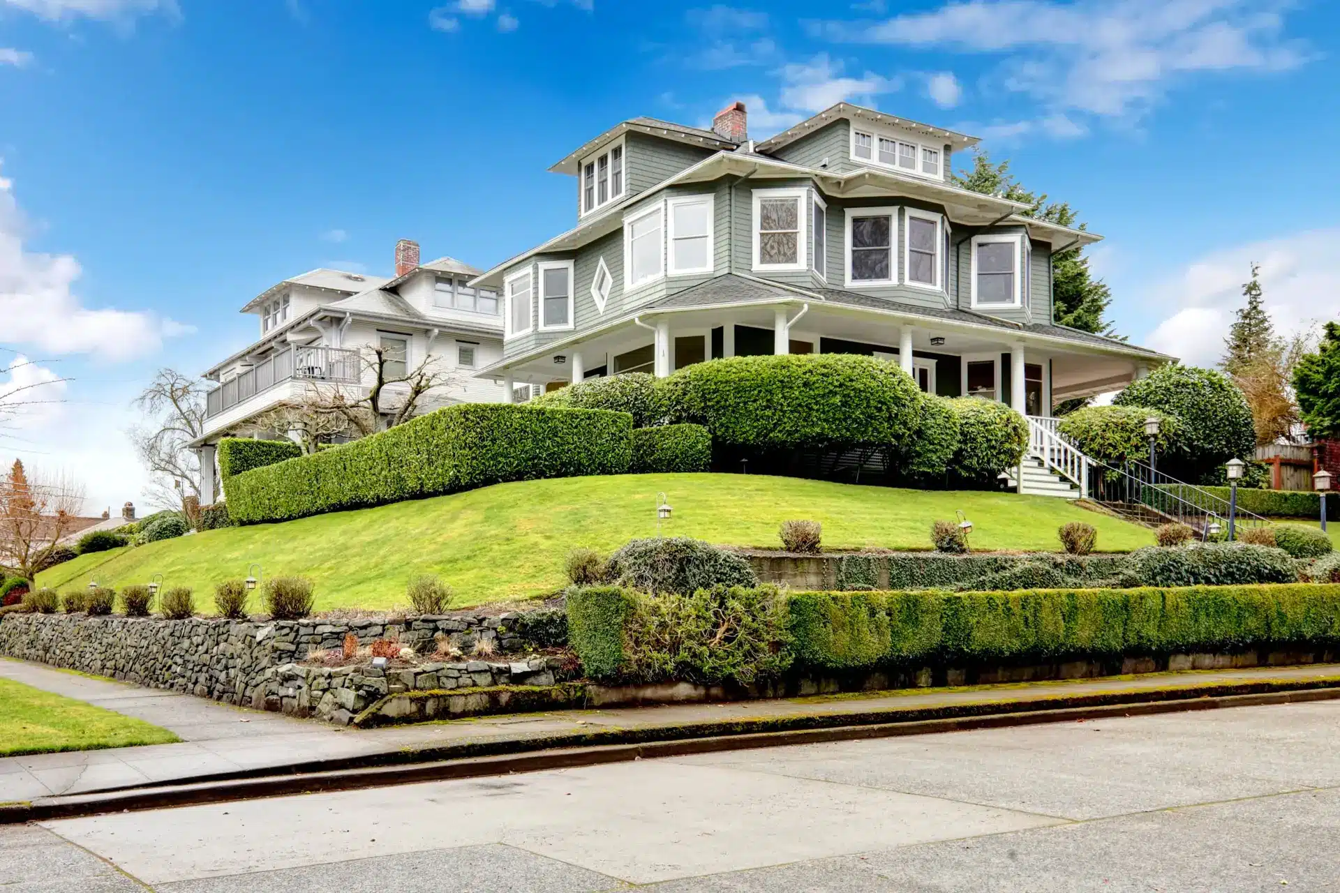 Large, two-story Victorian-style house with gray siding, white trim, and a wraparound porch showcases window trends 2024, surrounded by neatly trimmed hedges, a manicured lawn, and a stone retaining wall under a blue sky with clouds.
