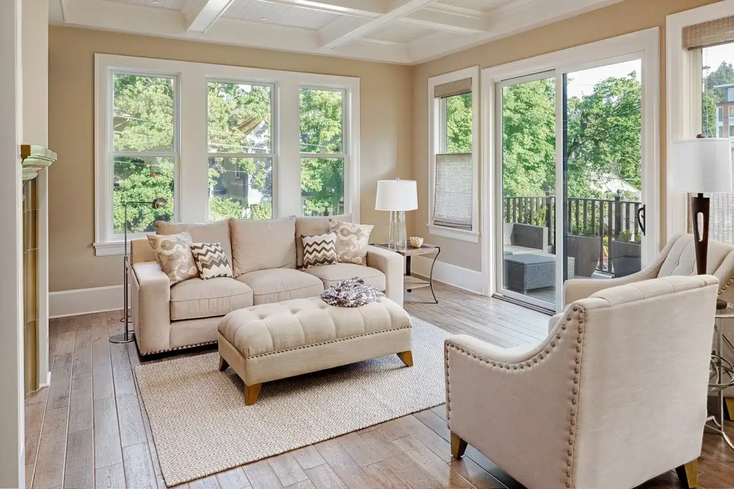 A bright living room with large windows showcasing a leafy view. It features a beige sofa, an ottoman, a cushioned armchair, and a floor lamp. A sliding glass door leads to a deck with outdoor seating. The room has hardwood floors and light walls. Vinyl Siding
