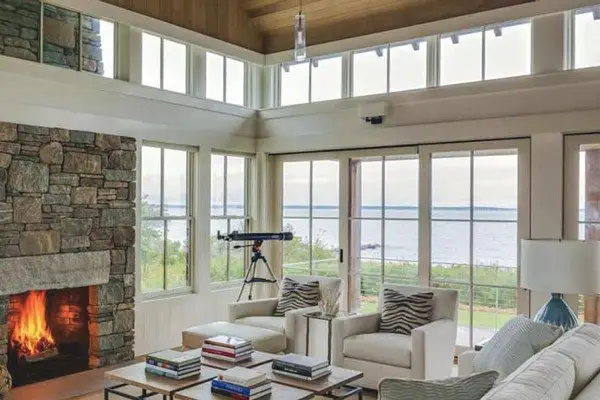 A bright living room with large vinyl windows overlooking water, a stone fireplace with a fire burning, two armchairs, a sofa, a coffee table with books, and a telescope facing the view—perfect for window shopping inspiration or your buying guide.
