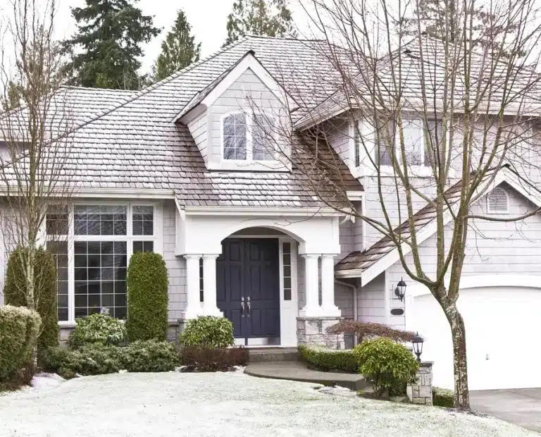 A light gray two-story house with a dark front door, arched window, and attached white garage. Trees and shrubs are in the front yard, and the ground is lightly dusted with snow—perfect for a Home Improvement Contractor’s next project.
