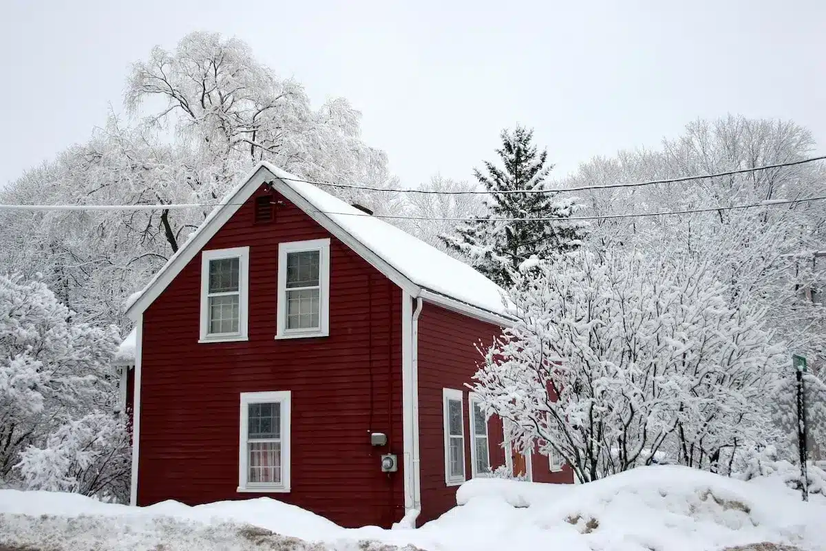 A small red house with white trim sits surrounded by snow-covered trees and bushes on a winter day. Snow piles up around the house, and the sky is overcast—an ideal reminder to winterize your home. Power lines run above the scene.