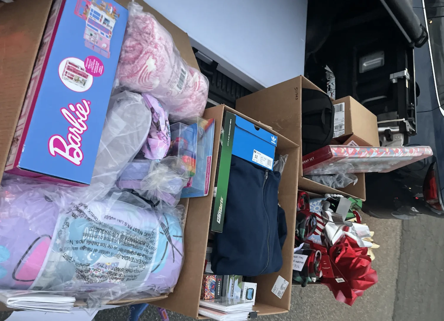 Several cardboard boxes in the trunk of a car are filled with wrapped gifts, toys including Barbie items, clothing, and holiday ribbon bows, suggesting Winter Wishes donations for Beverly Bootstraps or holiday gift preparations.