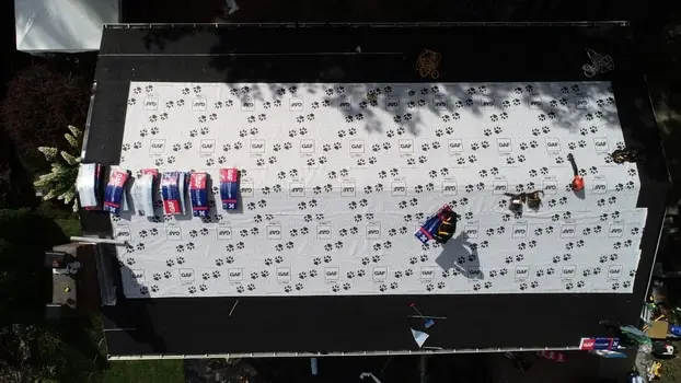 Aerial view of a rectangular roof being prepared for new shingles in Woburn, MA, with Superior Protection from the Elements. Roofing materials and tools are scattered on underlayment as workers operate, while tree shadows stretch across the surface.