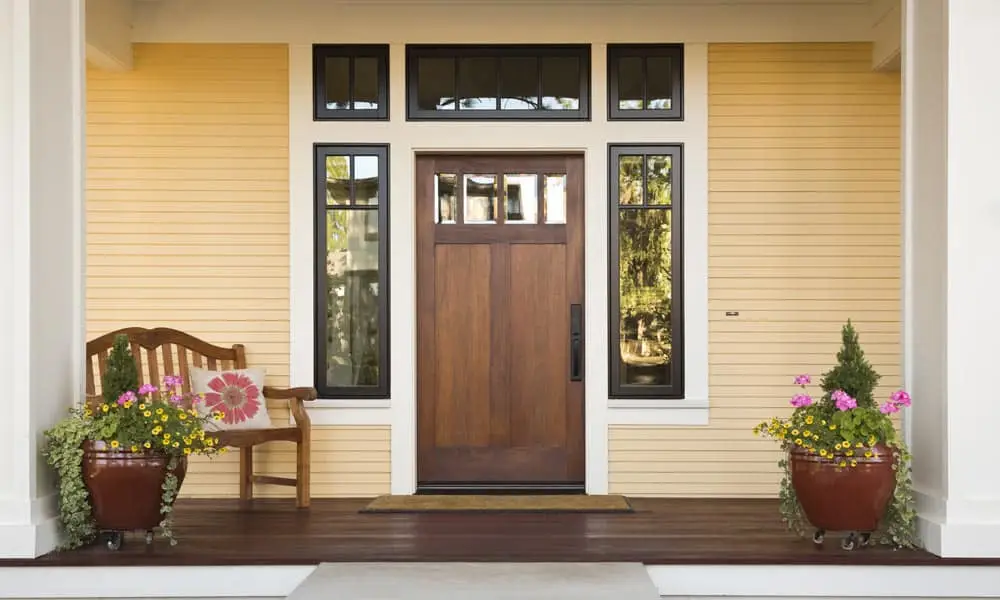 A wooden front door with glass panels is centered on a yellow house with white trim, adding curb appeal. Two large flowerpots with colorful flowers and a small bench with a floral cushion decorate the porch.