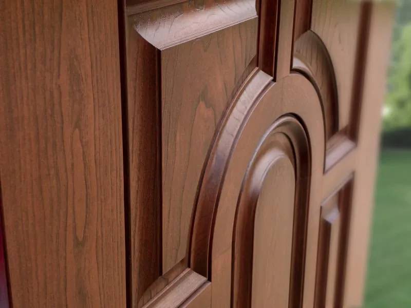 Close-up of a wooden door featuring intricate carvings and a natural grain pattern. The design includes geometric and curved elements, showcasing the craftsmanship. The background is blurred green foliage, suggesting an outdoor setting.