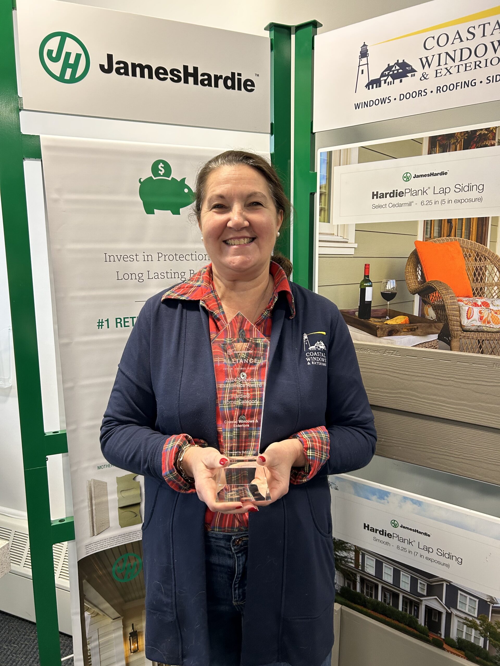 A smiling woman in a plaid shirt and blue jacket holds a small glass award. She stands proudly in front of James Hardie and Coastal Windows & Exteriors displays as a three-time James Hardie Service Excellence Award winner.