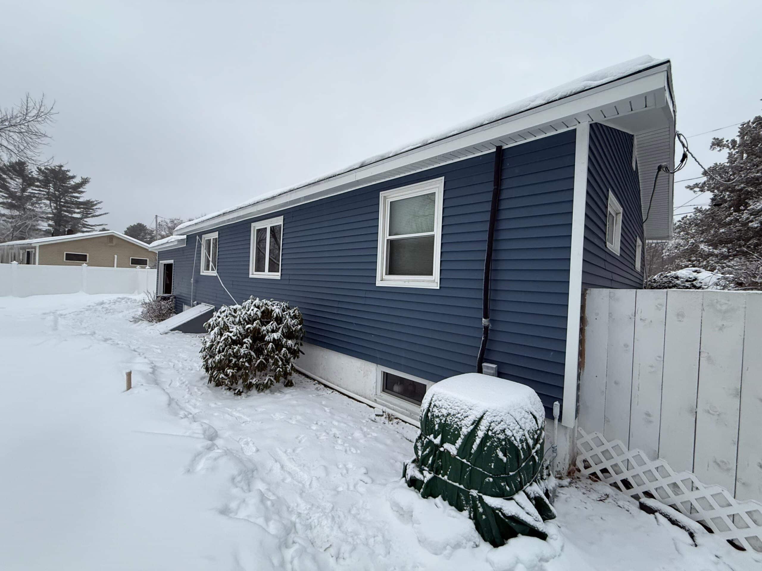 A blue house with white trim sits in a snowy yard. Shrubs and a green utility cover are partially covered in snow. A white fence and neighboring homes are visible in the background under a cloudy sky.