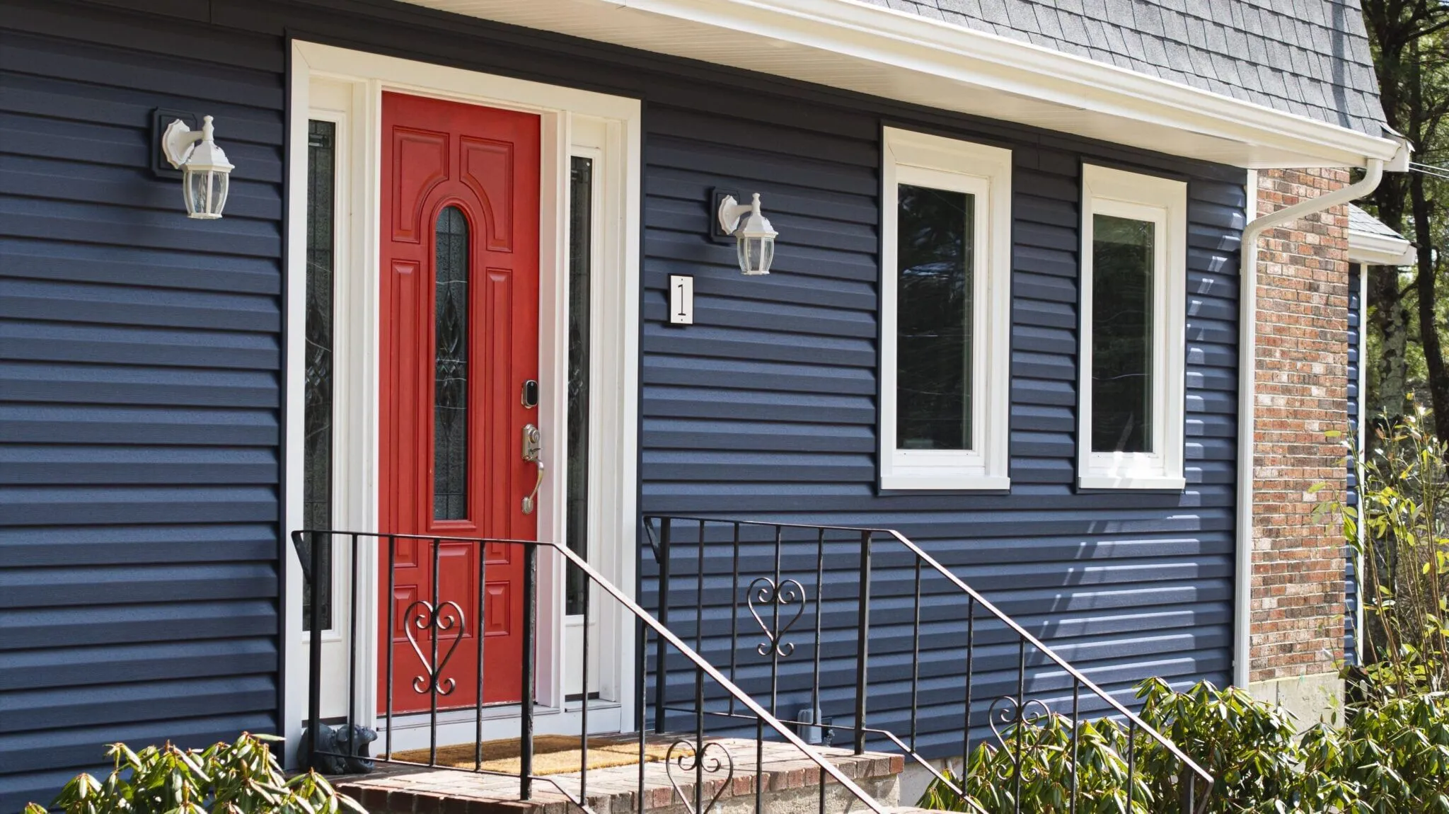 A bold red steel entry door creates a striking focal point against dark blue siding and white trim. Replacing an outdated front door is one of the most practical high-roi exterior upgrades for improving security, energy efficiency, and curb appeal.