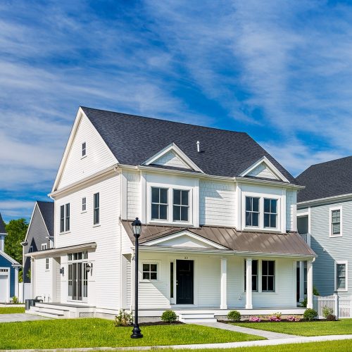 A modern two-story white house with vinyl siding in Rowley MA, featuring a gray roof, covered front porch, and neatly landscaped lawn under a blue sky with wispy clouds. A black lamppost stands near the walkway.